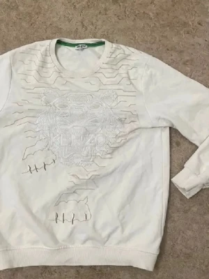 Vit sweatshirt från Kenzo med tiger - Säljer en vit sweatshirt från Kenzo med broderad tiger och grafiska linjer på framsidan. Tröjan har rund halsringning, ribbade muddar och är tillverkad i mjukt bomullsmaterial. Perfekt för dig som gillar stilrena designer med coola detaljer.