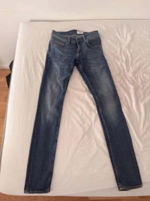 Blå slim fit jeans från Tiger of Sweden - Säljer ett par blå slim fit jeans från Nudie Jeans med klassisk femficksdesign och snygga slitningar på låren. Jeansen har kontrastsömmar och en brun patch bak i midjan. Materialet är denim med stretch för extra komfort. Använd bara fåtal gången, nypris ca 1400kr