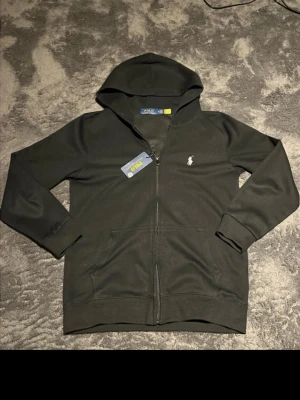 Svart hoodie från Polo Ralph Lauren - Snygg svart hoodie från Polo Ralph Lauren med dragkedja framtill och klassisk huva. Den har det ikoniska vita Polo-logotrycket på bröstet och ribbade muddar vid ärmar och nederkant. Tillverkad i mjukt bomullsmaterial för extra komfort. Ända defekten är att jag tagit ut snörerna till luvan och hittar tyvärr inte dom nu! Prislappen sitter tyvärr inte kvar, men den är ändå nästan helt oanvänd. Hör av er vid eventuella frågor 😊