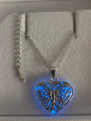Halsband med hjärthänge och fjäril - Unikt handgjort halsband med silverfärgad kedja i rostfritt stål och ett hjärtformat hänge med detaljerad fjäril. Hänget lyser blått i mörker efter att vanligt dagsljus eller solljus har nått hänget en stund. Bär halsbandet som vanligt så sköter sig effekten själv. Lyser vanligtvis i flera timmar & kan lysa upp till 12 timmar. Perfekt för dig som gillar smycken med lite extra wow-faktor och snygga detaljer.
