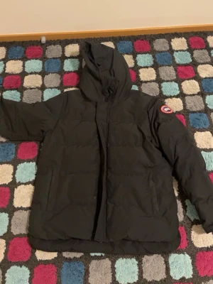 Svart dunjacka från Canada Goose - Säljer en svart dunjacka från Canada Goose med huva och klassisk logotyp på ärmen. Jackan har en rak passform, två sidofickor och dragkedja med vindslå. Perfekt för kalla vinterdagar och riktigt varm. Pris går att diskutera