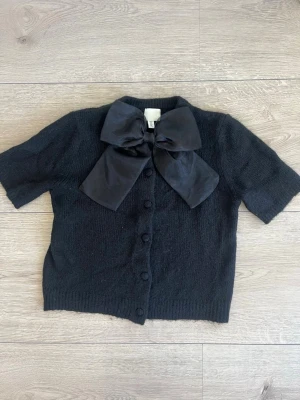 Svart kofta med stor rosett från H&M - Cool svart kofta från H&M med korta ärmar och knappar framtill. Den har en stor, elegant rosett i satinmaterial vid halsen som verkligen sticker ut. Perfekt för dig som vill ha något unikt och trendigt i garderoben. Den är i storlek M men passar i S, då den är lite liten i storlek.