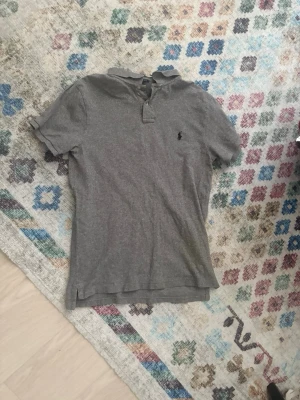 Grå pikétröja från Polo Ralph Lauren - Säljer en grå pikétröja från Polo Ralph Lauren med klassisk krage och knappar framtill. Tröjan har korta ärmar och slim fit-passform. Ikonisk logga broderad på bröstet. Perfekt för en avslappnad och stilren look.