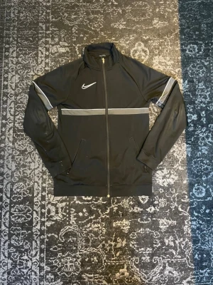 Svart Nike zip up tröja - Svart zip up tröja från Nike med hel dragkedja, ståkrage och vita detaljer över bröstet och armarna. Tröjan har två dragkedjefickor framtill och är tillverkad i ett mjukt, stretchigt material som passar perfekt till träning eller vardags outfit.