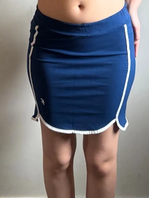 Blå sportig kortkjol med vita detaljer - Snygg blå kortkjol med vita kanter och sportig känsla. Kjolen har en liten slits på sidorna och elastisk midja för skön passform. Perfekt för en avslappnad och trendig look.