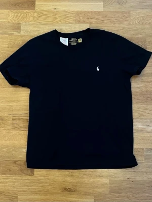 Svart t-shirt från Polo Ralph Lauren - En klassisk svart t-shirt från Polo Ralph Lauren med korta ärmar och rund hals. T-shirten har en liten vit broderad logotyp på bröstet och är tillverkad i mjuk bomull. Perfekt för dig som gillar stilrena basics.