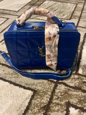 Blå YSL handväska med scarfdetalj - Snygg blå handväska från YSL i glansigt quiltat material med guldigt logotypemblem framtill. Väskan har både axelrem och handtag, där handtaget är dekorerat med en söt mönstrad scarf. Perfekt för att lyfta din outfit med en färgklick.