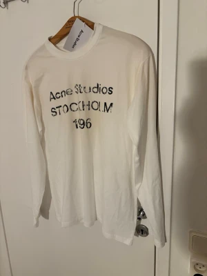 Vit långärmad topp från Acne Studios - Vit långärmad topp från Acne Studios med svart texttryck på bröstet där det står 'Acne Studios STOCKHOLM 196'. T-shirten har en klassisk rund halsringning och är gjord i mjuk bomull. Perfekt för dig som gillar minimalistisk stil och ikoniska märken.