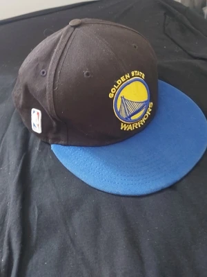 Golden State Warriors keps - Snygg keps med Golden State Warriors-logga broderad på framsidan. Kepsen är svart med blått flatbrätte och har NBA-märke på sidan. Perfekt för dig som vill visa ditt stöd för laget och ha en cool accessoar till din outfit.