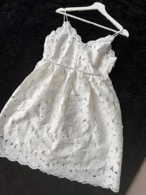 Vit spetsklänning med blommönster - Söt vit kortklänning med tunna axelband och vacker spets med blommigt mönster. Klänningen har en scallop-kant upptill och nertill samt fina broderade detaljer. Perfekt för sommarens alla tillfällen och ger en romantisk vibe.Passar till Studen och Brud😊 . Storleken är 40 på lappen men känns som 38 . Den är helt ny mmed prislapp . 