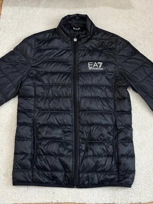 Dunjacka från EA7 Emporio Armani - Snygg svart pufferjacka från EA7 Emporio Armani med diskret glansig finish och logga på bröstet. Jackan har ståkrage, dragkedja framtill och två sidofickor.                                                                        Perfekt nu på våren och ger en clean streetwear-look. QR-kod till Certilogo (äkthetskontroll)                                                                             Nypris: 2400kr