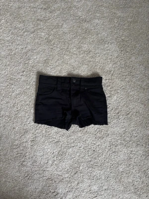 Svarta denimshorts med rå kant - Säljer ett par svarta denimshorts med rå, fransig kant och klassisk femficksdesign. Shortsens midja är normal och de har knapp och dragkedja framtill. Perfekta för sommarens alla äventyr!