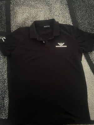 Svart pikétröja från Emporio Armani - Säljer en svart pikétröja från Emporio Armani med klassisk krage och två knappar. Tröjan har ett vitt logotryck på bröstet och är tillverkad i ett mjukt material som känns bekvämt. Perfekt för dig som gillar stilrena och exklusiva plagg.