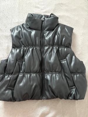 Svart glansig väst med dragkedja - Snygg svart väst i glansigt material med hög krage och dragkedja framtill. Västen har två snedställda fickor och quiltade sömmar som ger en puffig look. Perfekt på våren och hösten när det är för varmt med jacka men för kallt för bara T-shirt!