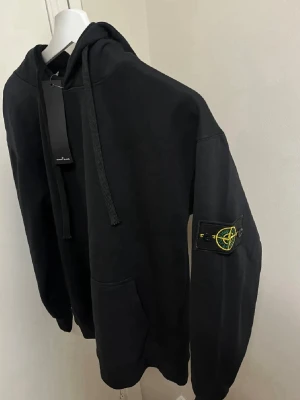 Svart hoodie från Stone Island - Snygg svart hoodie från Stone Island med klassisk patch på vänster ärm. Hoodien har stor huva, breda dragsnören och en rymlig magficka. Tillverkad i mjukt bomullsmaterial som känns skönt mot huden. Perfekt för en avslappnad och trendig streetwear-look. Pris kan diskuteras
