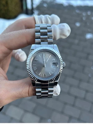 Silverfärgad klocka med datumfönster - Stilren klocka i silver med klassisk länkarmband och räfflad bezel. Urtavlan är också silverfärgad med romerska siffror och datumfönster vid klockan tre. Klockan har ett automatiskt urverk som syns genom baksidan. Extra länkar medföljer.