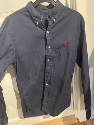 Mörkblå skjorta från Polo Ralph Lauren - Säljer en klassisk mörkblå skjorta från Polo Ralph Lauren med lång ärm och knappar framtill. Skjortan har den ikoniska röda broderade loggan på bröstet och är tillverkad i bomull. Perfekt för dig som gillar stilrena plagg med snygga detaljer.