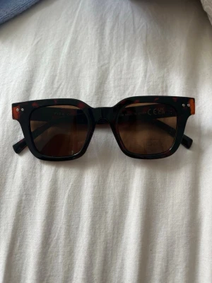 Bruna solglasögon från Pier One - Snygga solglasögon från Pier One med breda, fyrkantiga bågar i brun och brunmelerad design. Glasen är mörka och ger ett coolt uttryck. Perfekt accessoar för att lyfta din stil under soliga dagar. Aldrig använda 