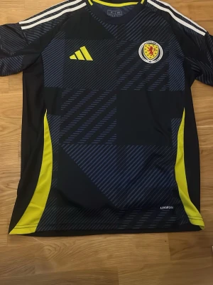 Skottland matchtröja Adidas blå/gul - Helt oanvänd Snygg replika matchtröja från Skottlands landslag i mörkblått med gula detaljer och vita ränder på axlarna. Adidas-logga och Skottlands klubbmärke på bröstet. Tillverkad i lätt och ventilerande material med Aeroready-teknologi. Perfekt för fotbollsträning eller match.
