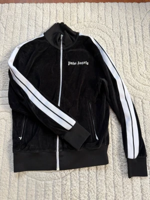 Svart träningsjacka Palm Angels - Snygg svart träningsjacka från Palm Angels i mjuk velour med vit logotyp på bröstet. Jackan har hel dragkedja framtill, ribbade muddar och två fickor med dragkedja. Perfekt för en sportig och trendig look.