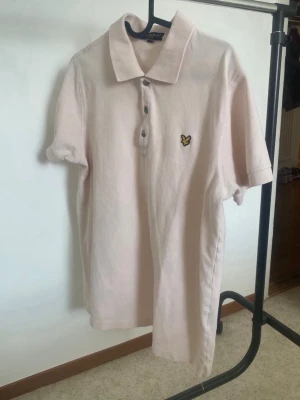 Ljusrosa pikéskjorta från Lyle & Scott - Säljer en ljusrosa pikéskjorta från Lyle & Scott med klassisk krage och knappar framtill. Skjortan har korta ärmar och ett broderat logomärke med gul detalj på bröstet. Perfekt för dig som gillar stilrena plagg med diskreta färger.
