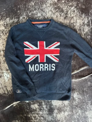 Morris mörkblå tröja med Union Jack - Mörkblå stickad tröja från Morris med Union Jack-motiv i rött, vitt och blått på bröstet samt Morris-tryck. Tröjan har ribbade muddar och rund halsringning. Perfekt för dig som gillar brittisk stil och vill ha något som sticker ut.