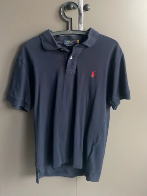 Marinblå pikétröja Polo Ralph Lauren - Marinblå pikétröja från Polo Ralph Lauren med klassisk krage, två knappar och röd broderad logga på bröstet. Tröjan är kortärmad och tillverkad i mjuk bomull som är skön att bära. Perfekt för dig som gillar en stilren och tidlös look. Passar perfekt under sommaren.               Hör gärna av dig vid frågor eller funderingar!  Mvh Melker