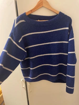 Blå stickad tröja med vita ränder - Mysig blå stickad tröja med vita horisontella ränder och rund hals. Tröjan har en avslappnad passform och långa ärmar. Perfekt att slänga på sig för en chill look. Materialet ser ut att vara mjukt och lite fluffigt, vilket gör den extra bekväm.