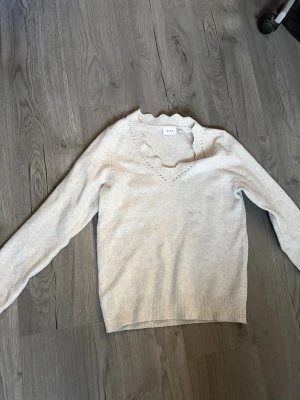 Beige stickad tröja med volangkant - Säljer en ljusbeige stickad tröja från Vila med lång ärm och v-ringning. Tröjan har en söt volangkant runt halsen och ribbade muddar vid ärmslut och nederkant. Perfekt för dig som gillar en enkel men ändå feminin stil. Jag säljer då den inte kommer till användning💕