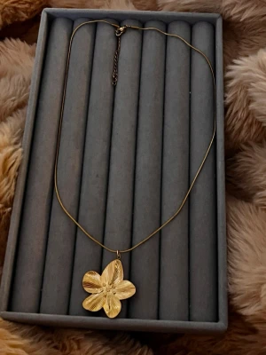 Guldigt halsband med blomma - Snyggt halsband i guldfärgad metall med ett stort blomhänge. Kedjan är tunn och smidig med justerbar längd och karbinlås. Blomman har fina detaljerade blad som ger ett elegant intryck. Perfekt accessoar för att lyfta din outfit.