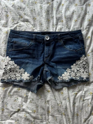 Blå jeansshorts med vita spetsblommor - Snygga blå jeansshorts med spetsdesign från Ellos som har fransig kant, fem fickor och knappgylf. Storleken har skavt bort i lappen men uppskattar de som en M 
