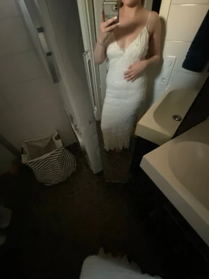 Vit långklänning med volangdetaljer - Säljer en elegant vit långklänning med tunna axelband och v-ringning. Klänningen har fina volangdetaljer och omlottkänsla upptill. Perfekt för sommarens alla tillfällen. Materialet känns lätt och luftigt.