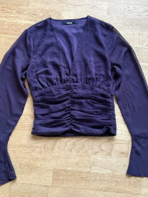 Lila blus med urringning från Bik Bok - Snygg plommonlila blus från Bik Bok med djup v-ringning och rynkade detaljer framtill. Blusen har långa ärmar och är tillverkad i ett tunt, lätt material som ger en elegant känsla. Perfekt för att skapa en trendig look.