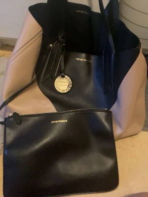 Emporio Armani shoppingväska svart/beige - Stilren shoppingväska från Emporio Armani i svart och beige konstläder. Väskan har två axelband, ett stort huvudfack och ett guldfärgat emblem med logga. Medföljer gör en matchande svart clutch med dragkedja, perfekt för småsaker.
