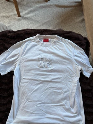 Vit HUGO t-shirt med 3D-logga - Säljer en clean vit t-shirt från HUGO med upphöjd 3D-logga på bröstet. Klassisk rund halsringning och korta ärmar. Tillverkad i mjuk bomull som känns skön mot huden. Perfekt för dig som gillar stilrena plagg med en twist.