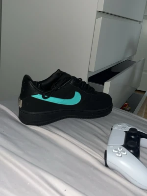 Nike Air Force 1 Tiffany  - Säljer ett par Nike Air Force 1 sneakers i svart läder med en fet turkos swoosh på sidorna. Klassisk låg modell med svarta skosnören och svart sula. Perfekta för dig som vill sticka ut med en clean och modern look. Nästan nya har använd du dem i 1-2 månader 