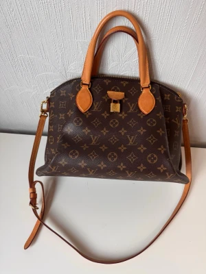 Louis Vuitton Monogram handväska - Snygg handväska från Louis Vuitton i klassiskt brunt monogrammönster med ljusbruna läderdetaljer. Väskan har både handtag och justerbar axelrem samt guldfärgade metalldetaljer. Tillverkad i canvas och läder, perfekt för dig som gillar lyxiga accessoarer.