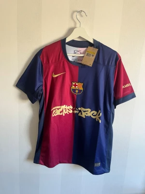 Barça x Travis Scott matchtröja Nike - Säljer en officiell FC Barcelona matchtröja i samarbete med Travis Scott och Nike. Tröjan är röd och marinblå med klubbmärke, Nike-logga och Travis Scott-grafik på bröstet. Tillverkad i lätt, ventilerande material och har korta ärmar.