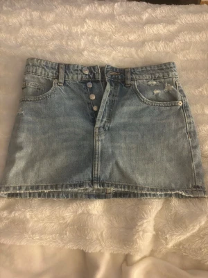 Ljusblå jeanskjol med knappar - Trendig ljusblå jeanskjol i kort modell med synliga knappar framtill och rå kant nedtill. Kjolen har klassiska fem fickor och bälteshällor. Perfekt till sommaren och enkel att matcha med olika toppar. Använd en gång, från Zara. Säljs då den är förliten💓