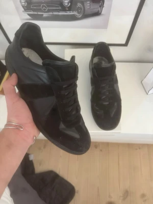 Svarta sneakers från Maison Margiela - Svarta sneakers från Maison Margiela med en mix av mocka och läder. Klassisk låg modell med svarta skosnören och diskret design. Perfekta för dig som gillar stilrena och exklusiva sneakers.