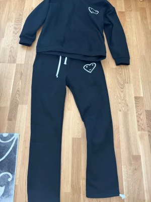 Svart loungewear-set med hjärtdetalj - Mysigt svart loungewear-set med sweatshirt och byxor. Båda plaggen har en vit broderad hjärtdetalj med ansikte. Byxorna har dragsko i midjan för skön passform. Perfekt för chill dagar hemma.