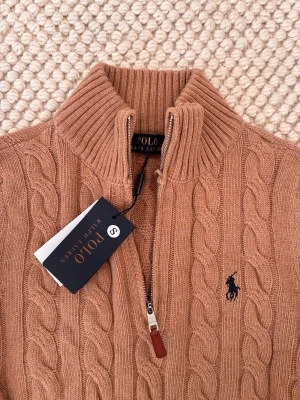 Beige kabelstickad tröja Polo Ralph Lauren - Säljer en beige kabelstickad tröja från Polo Ralph Lauren med halv dragkedja och hög krage. Tröjan har ribbade muddar och det klassiska mörkblå logobroderiet på bröstet. Perfekt för dig som gillar stilren och tidlös design.