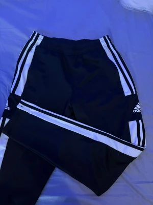 Svarta Adidas träningsbyxor med vita ränder - Svarta träningsbyxor/överdragsbyxor från Adidas med klassiska vita ränder längs sidorna och dragkedja vid bensluten. Byxorna har resår i midjan och Adidas-logga på låret. Perfekta för en sportig och avslappnad stil.