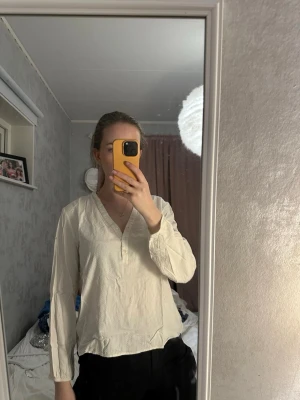 Krämvit blus med v-ringning - Stilren krämvit blus med v-ringning och knappar framtill. Blusen har långa ärmar och är tillverkad i ett lätt, något skrynkligt tyg som ger en avslappnad look. Perfekt att matcha med både jeans och kjol för en trendig vardagsstil.
