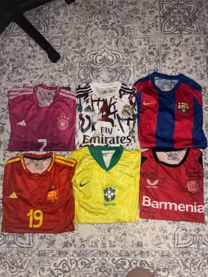 6 fotbollströjor från Adidas och Nike - Säljer ett paket med sex fotbollströjor: Tysklands rosa Adidas-tröja, Real Madrids vit/svart Adidas-tröja, Barcelonas blå/röd Nike-tröja, Spaniens röd/gul Adidas-tröja, Brasiliens gul/grön Nike-tröja och Bayer Leverkusens röd Castore-tröja. Perfekt för fotbollsfans!