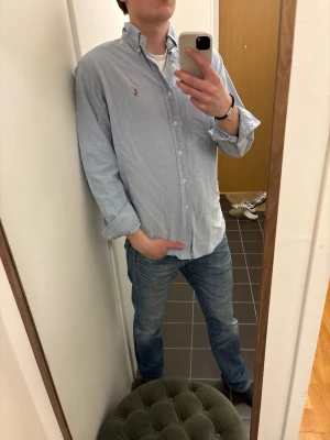 Ljusblå skjorta från Ralph Lauren - Klassisk ljusblå skjorta från Ralph Lauren med button-down krage och broderad logga på bröstet. Skjortan har långa ärmar och knäppning framtill. Perfekt för en stilren och avslappnad look.