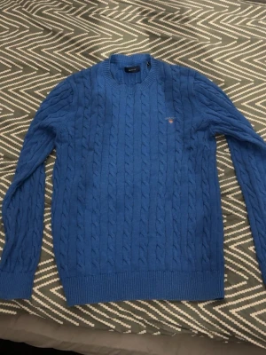 Blå kabelstickad tröja från Gant - Snygg blå kabelstickad tröja från Gant med rund halsringning och diskret logga broderad på bröstet. Tröjan har långa ärmar och ribbade muddar vid ärmslut och nederkant. Perfekt för lager på lager och en klassisk stil.