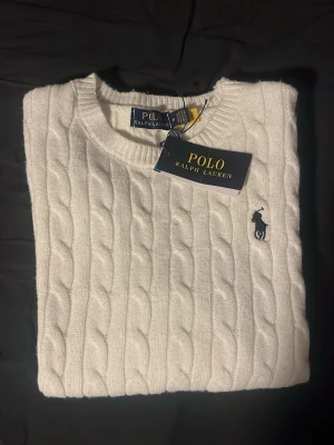 Vit kabelstickad tröja Polo Ralph Lauren - Vit kabelstickad tröja från Polo Ralph Lauren i storlek M. Klassisk design med rund hals och broderad logga på bröstet. Tröjan har långa ärmar och ribbade muddar vid hals, ärmslut och nederkant. Perfekt för dig som gillar stilrena och tidlösa plagg. Priset kan diskuteras. 