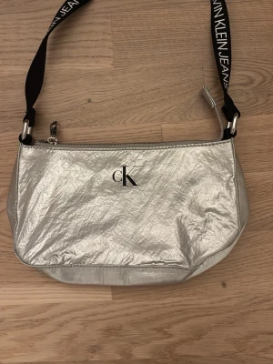 Silvrig handväska från Calvin Klein - Snygg silvrig handväska från Calvin Klein Jeans med svart axelrem där märkesnamnet står tryckt i vitt. Väskan har ett stilrent cK-logo framtill och dragkedja upptill. Materialet är glansigt och ger en modern look. Perfekt accessoar för att lyfta din outfit.