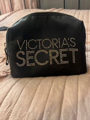 Svart necessär Victoria's Secret - Snygg svart necessär från Victoria's Secret med glittrande nitar som bildar loggan på framsidan. Väskan har en mjuk, rektangulär form och dragkedja upptill. Perfekt för smink eller småprylar när du är på språng.